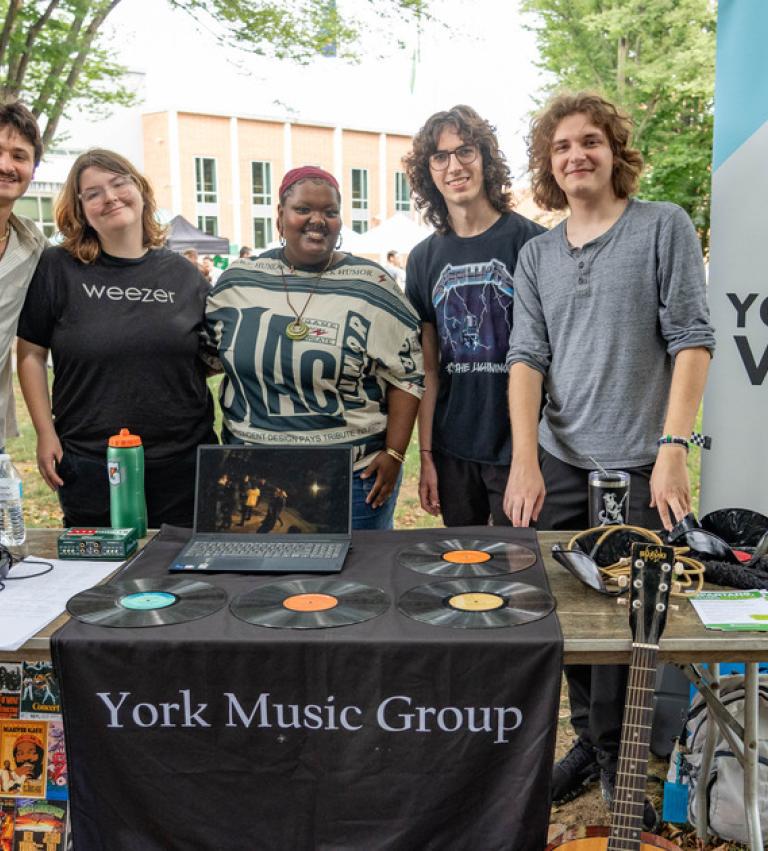Five students stand behind a decorated table that says York Music Group at the Summer Involvement Fair