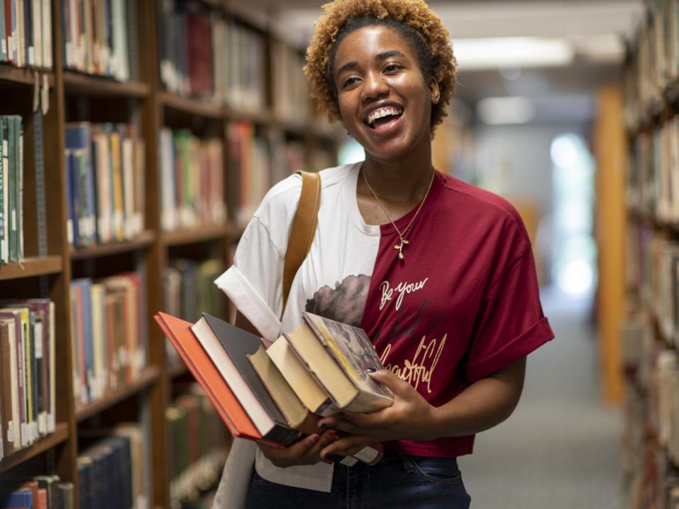 A person smiles brightly while holding books in a library. Full bookshelves are visible on either side and there is a window in the background.