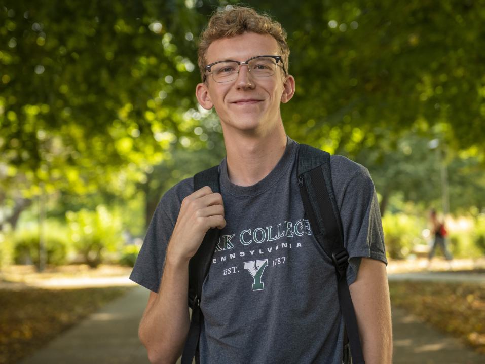 Student in a York College tee shirt and glasses holds the strap of his backpack while walking on campus on a sunny day.