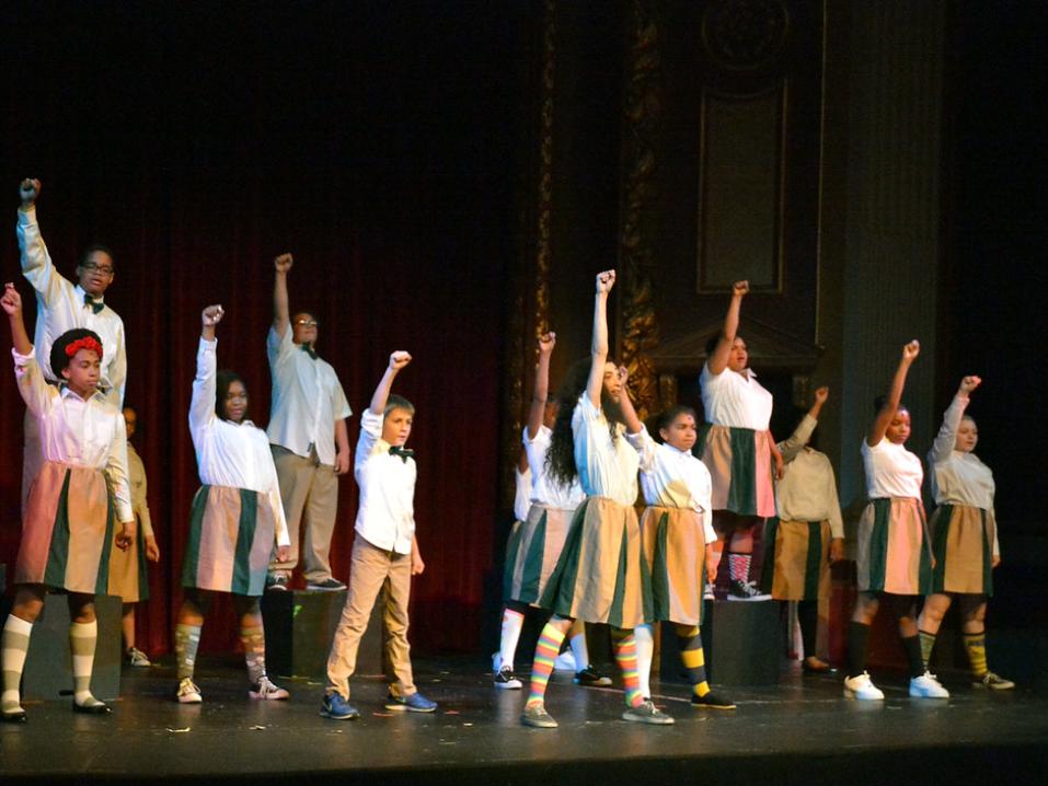 A dark stage with a group of children in costume, each with one arm raised in the air.