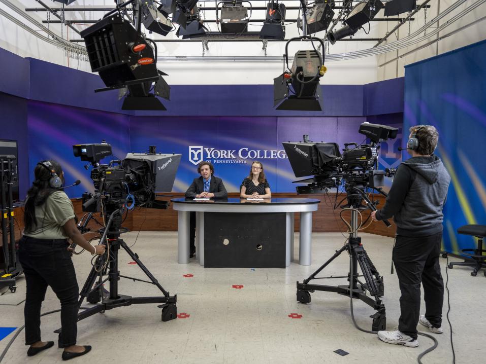 A wide view of a tv studio with large studio cameras, lights overhead, and two people sitting at a desk with the words "York College" behind them