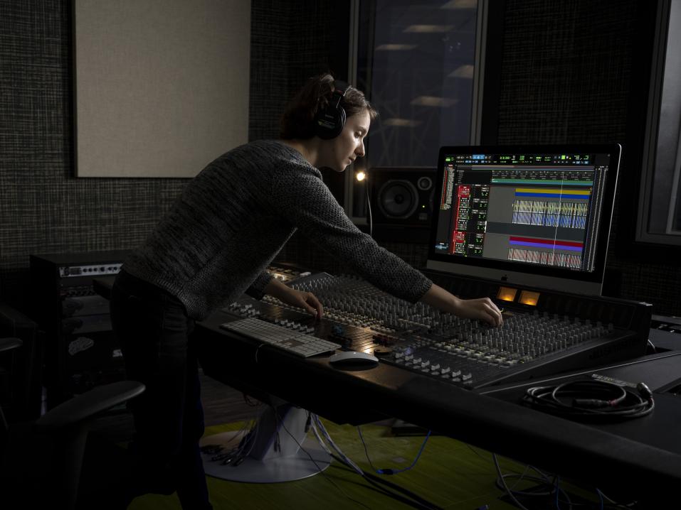 A student wearing headphones in front of a large audio board and computer monitor
