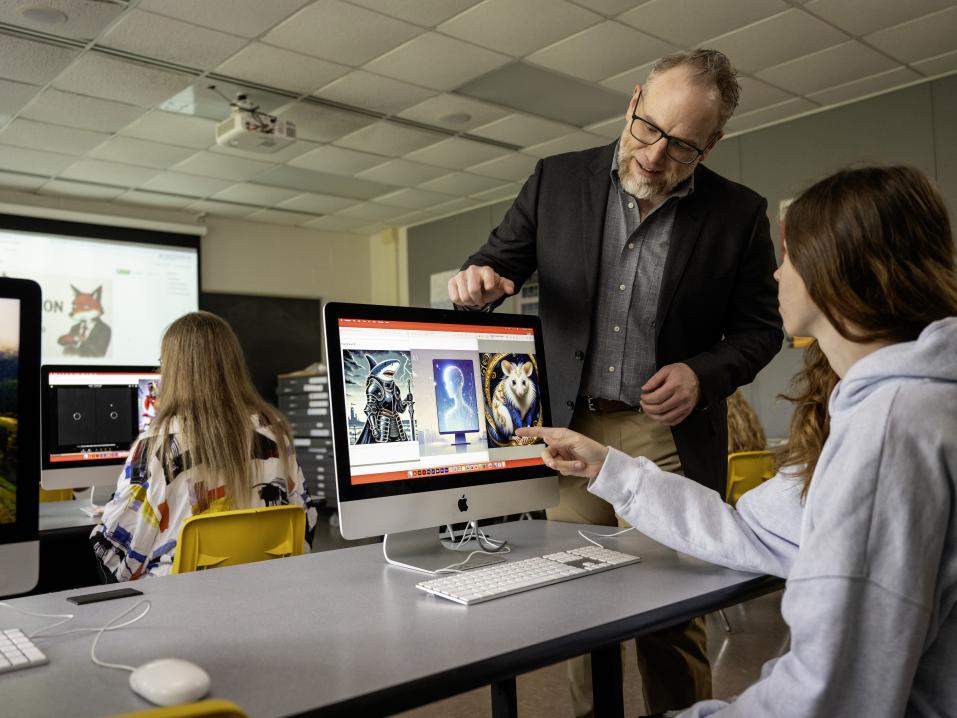 A classroom space with several computers featuring digital art. A professor looks over a student's work on a computer.