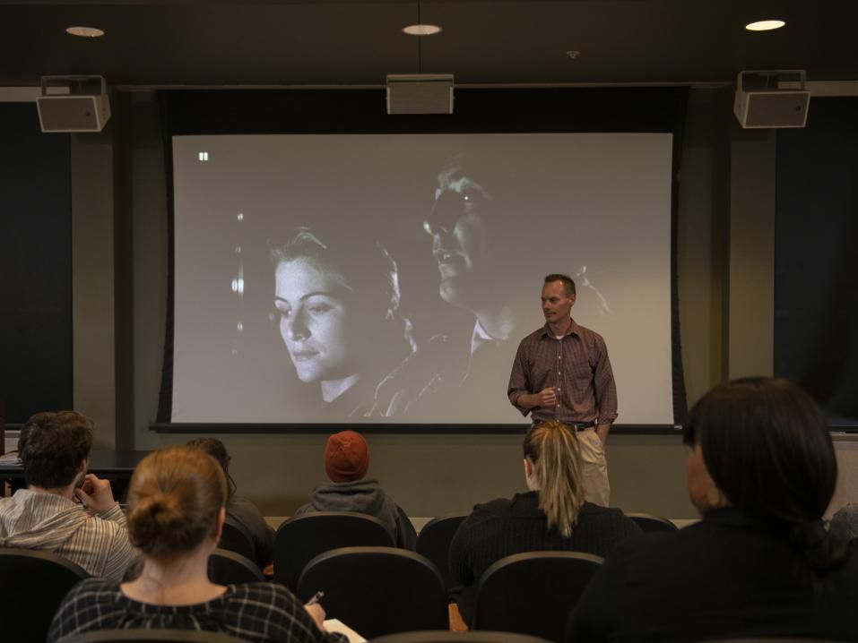 A man stands in front of a movie screen with a black and white image. The backs of audience members can be seen. 