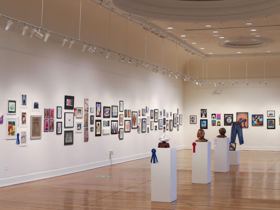 A large open art gallery with white walls and warm wood floors. The walls are covered in artwork.