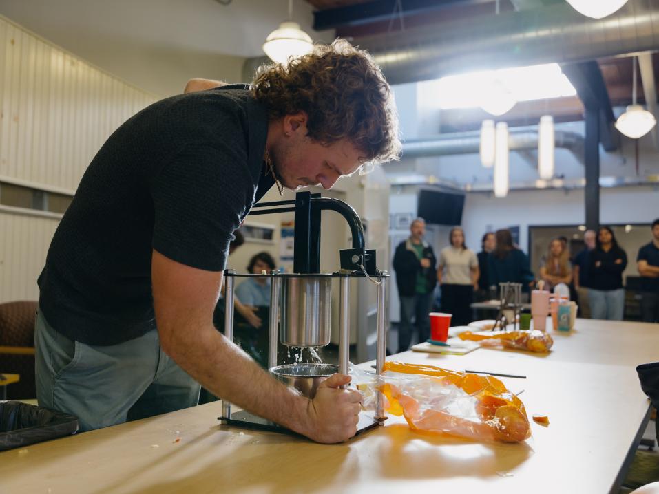 A student operates a manual apple press at a table, pressing chopped apples into a container. People watches in the background inside an engineering lab.