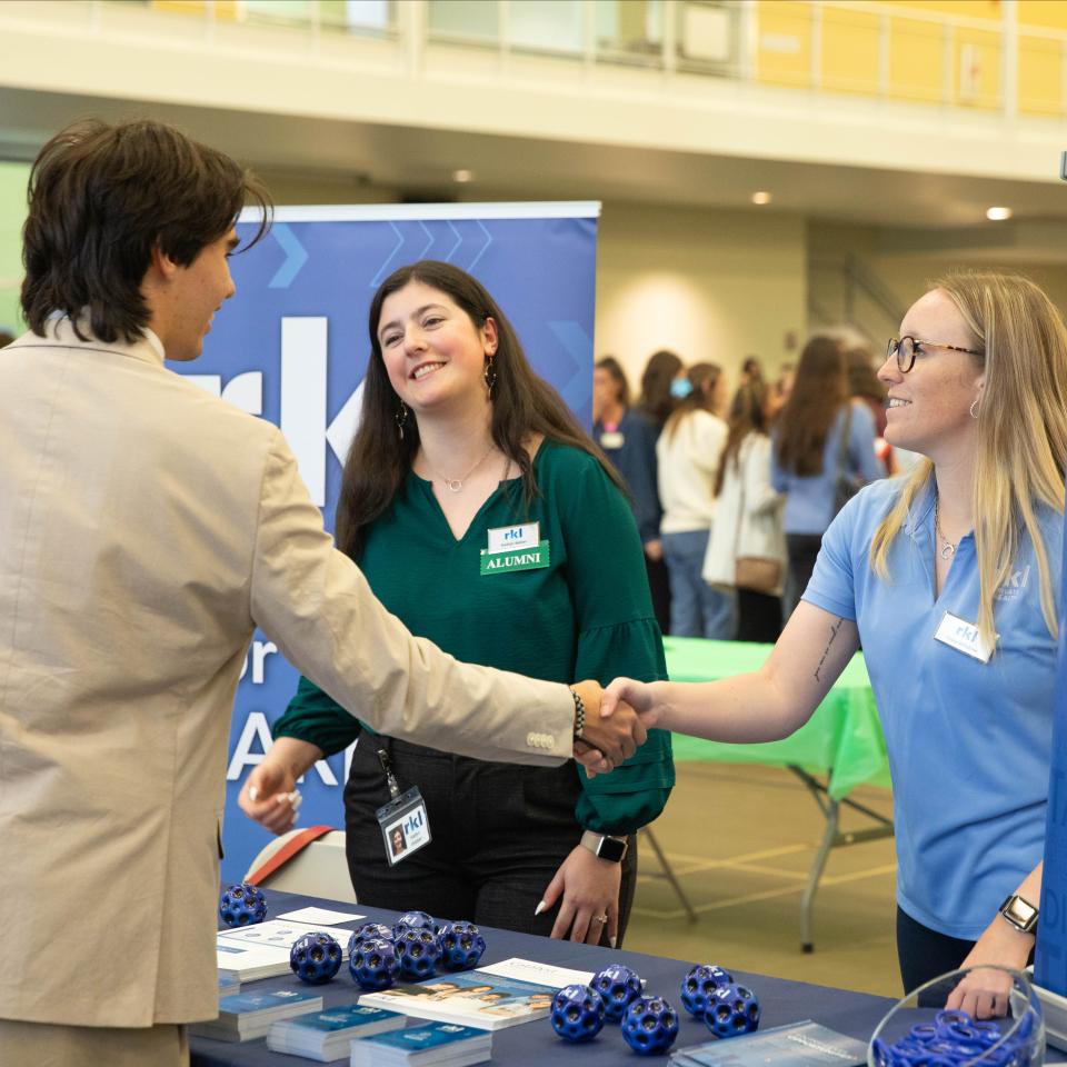 Two professionals shake hands at an expo event while a third smiles.