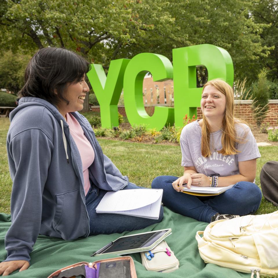 Three female students sit outside on a blanket with the YCP letters behind them.