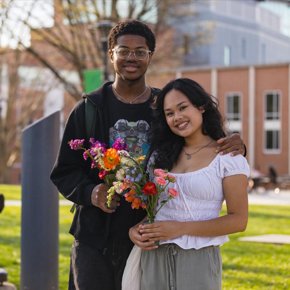 Two students pose and smile outside holding flowers with a brick building behind them.