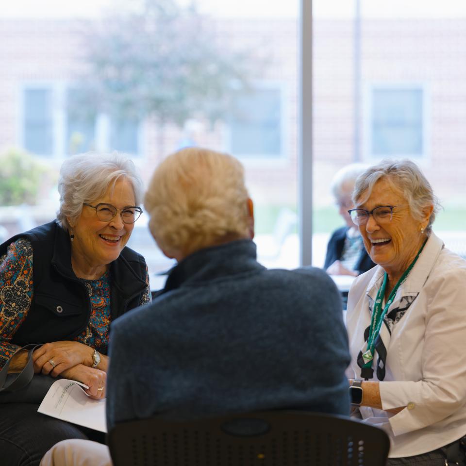 Three women sit at a table smiling and talking with a large window behind them.
