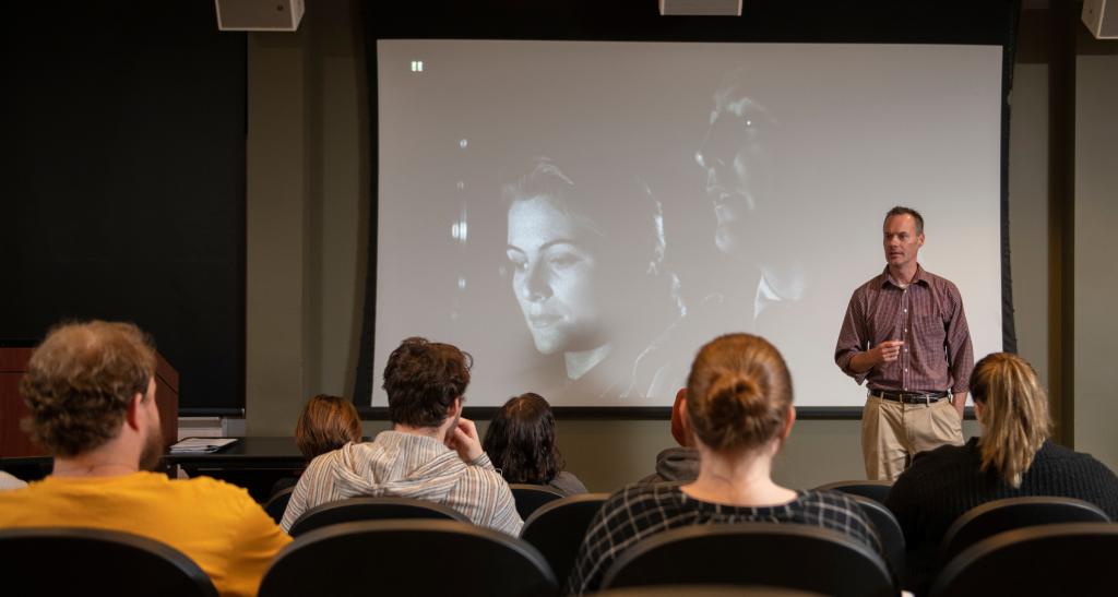 A professor stands at the front of a film screening room, speaking to the seated crowd as a black and while film plays on the projection screen behind him.