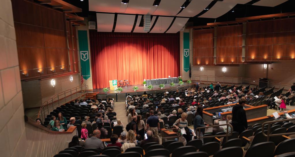 A wide view of a theater full of people with red curtains and green banners.