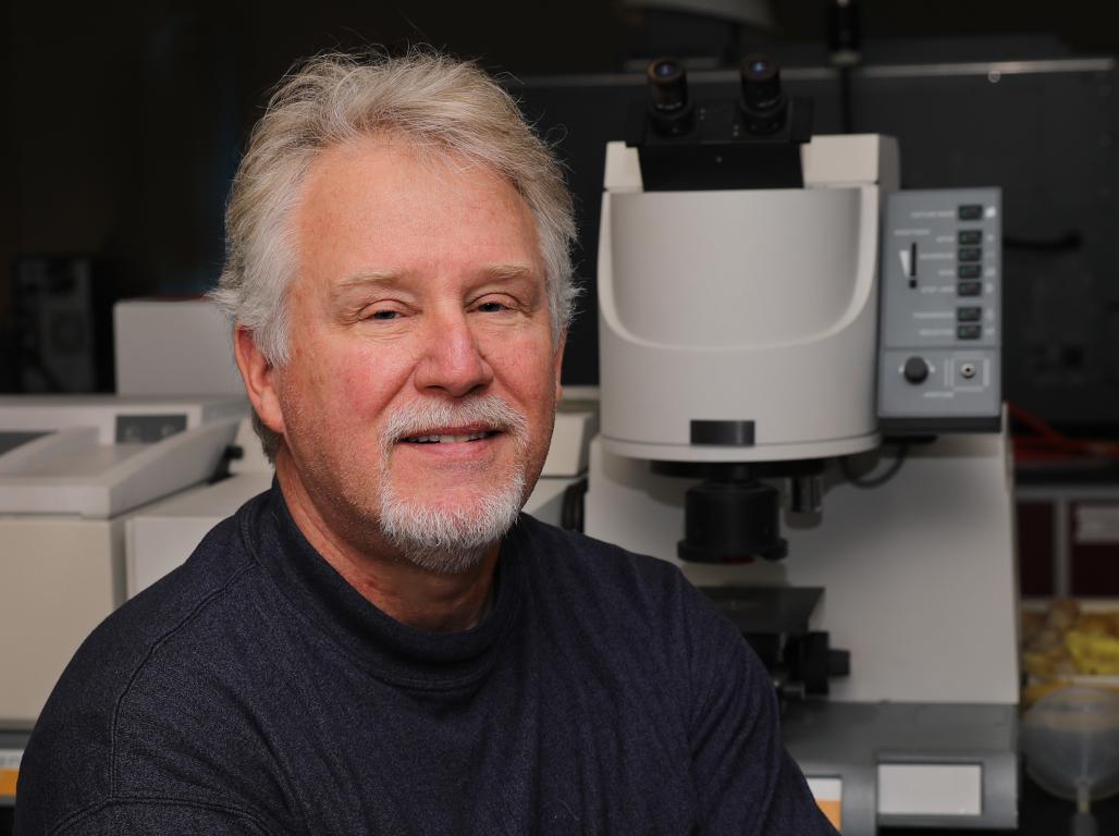Dr. Greg Foy smiles at the camera in a lab setting with chemistry equipment visible in the background.