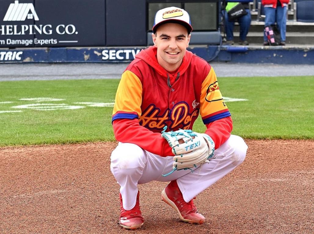 A baseball player wearing a red, white, and yellow uniform crouches on a baseball field with a catcher's mitt, smiling at the camera.