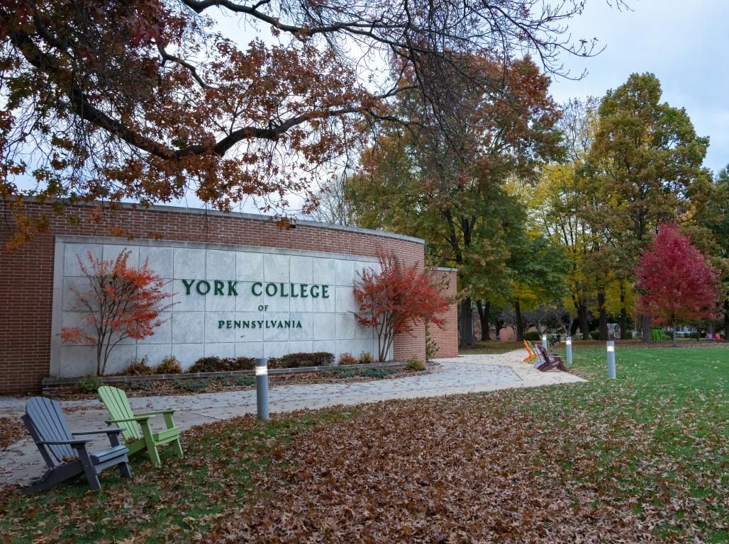 An exterior brick wall with "York College of Pennsylvania" signage on a fall day. A sidewalk, trees with autumn foliage, and outdoor chairs are visible.