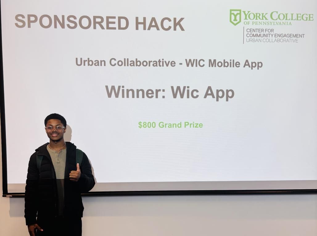 Computer Science Major Chibudom Onyejesi '27 standing in front of a projector screen indicating that his project won the 2025 YCP Hackathon.