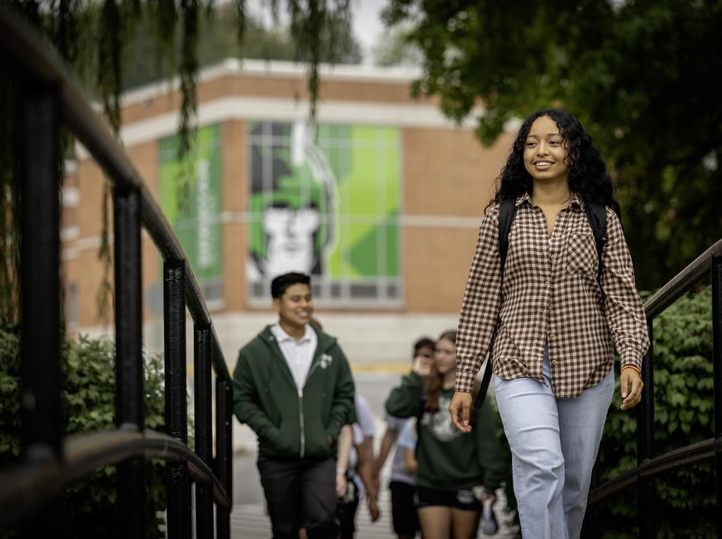 College students walking across an outdoor bridge. Trees and a brick building with a large green decal are visible in the background.