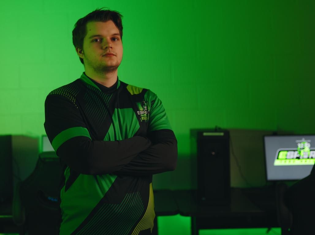 Person wearing a green and black ESports shirt poses with arms crossed in a dimly lit green room with computers visible in the background.