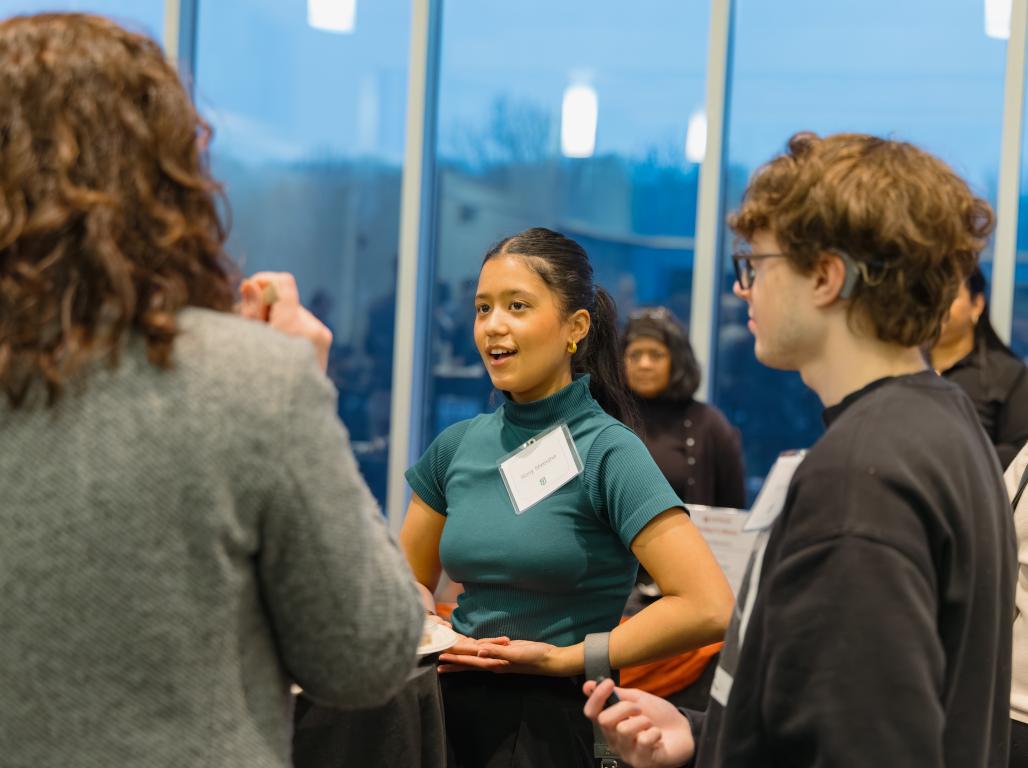 Three people speaking to each other in a crowded room with large windows behind them.
