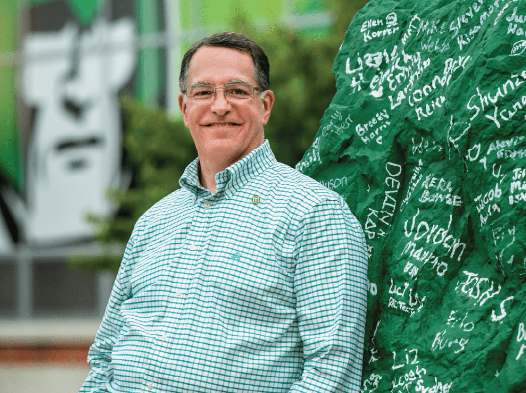 Dr. Burns, president of York College, leaning against a rock that's painted green with names handwritten in white on the surface.