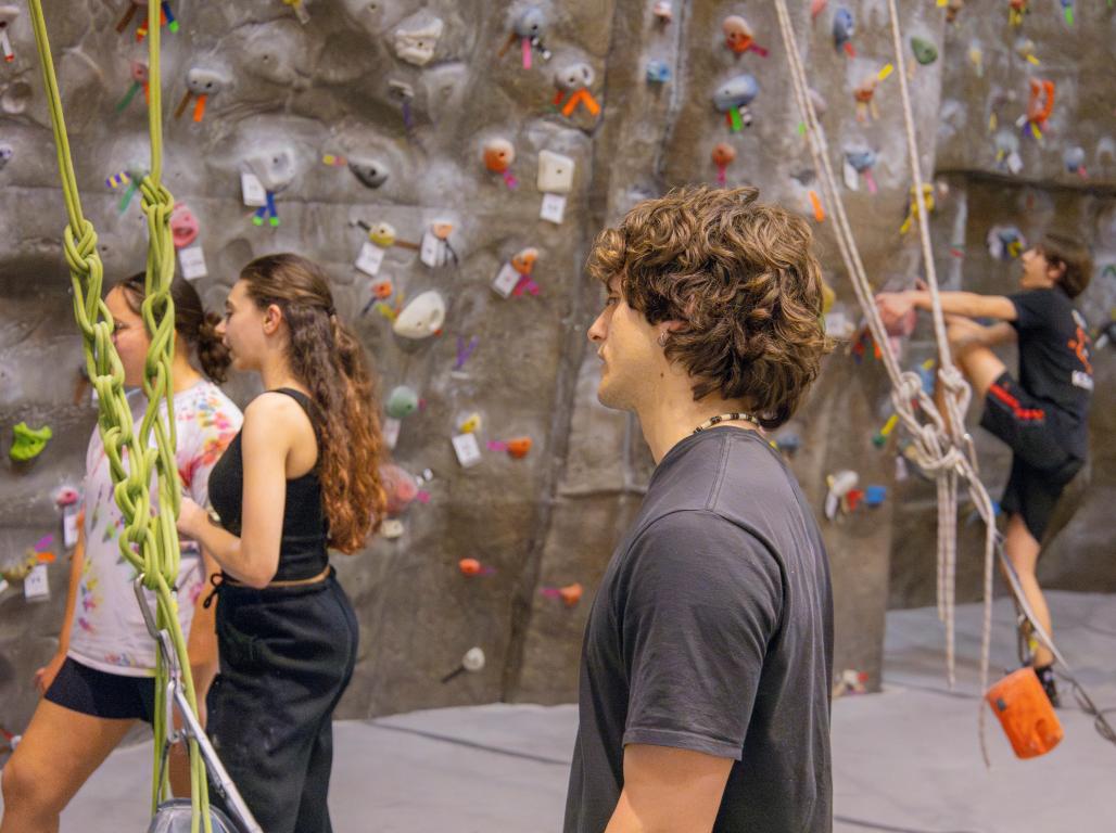A group of people at an indoor rock climbing wall, some are looking at the wall, while an active climber is visible in the background.