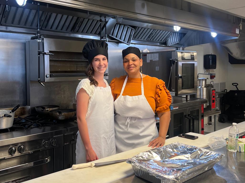 Two women stand in an industrial kitchen wearing aprons and hats.