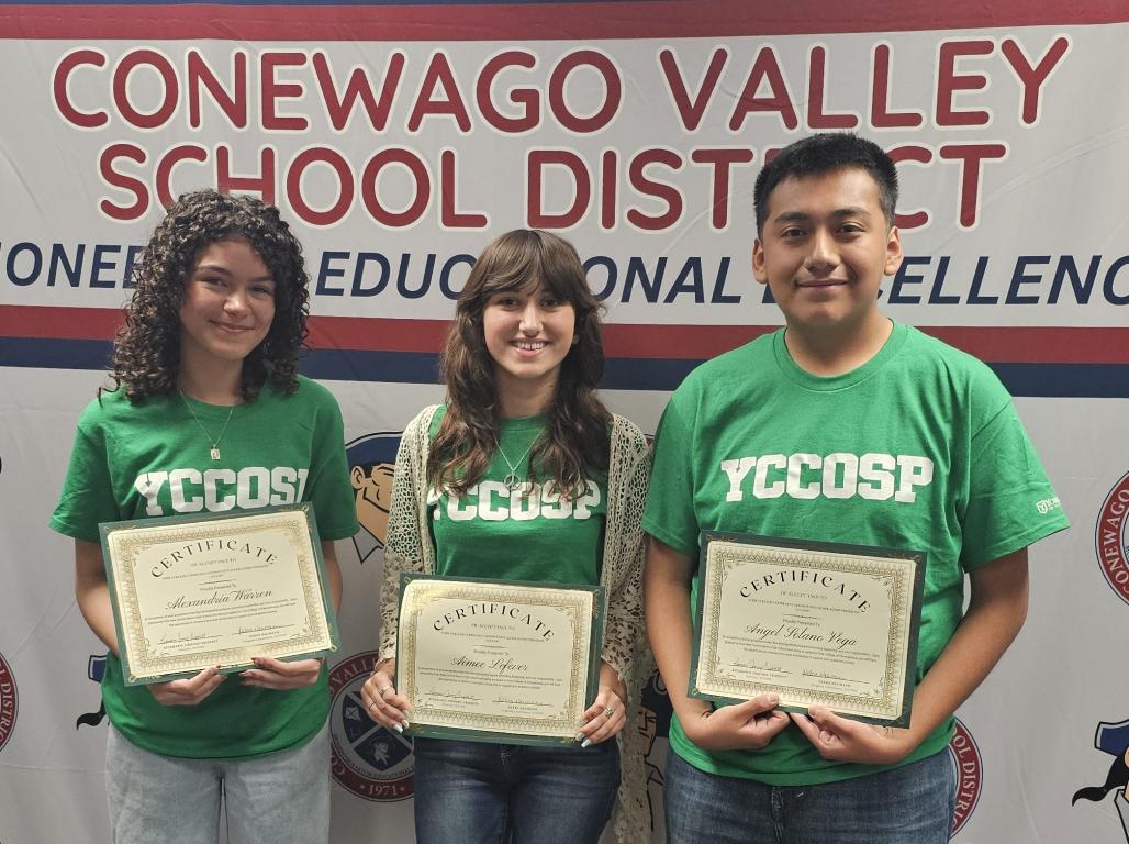 Three students in green tee shirts with "YCCOSP" in green lettering smile at the camera while holding certificates. Behind them is a backdrop that says "Conewago Valley School District" with logos in a repeating pattern.