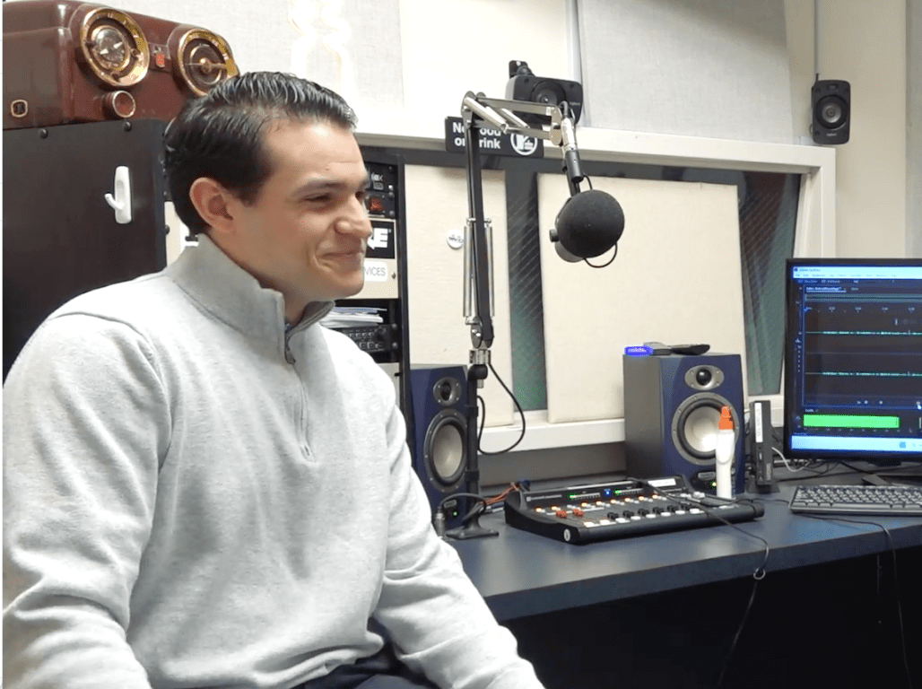 A student and a professor sit in a podcast room with a computer and microphones between them.