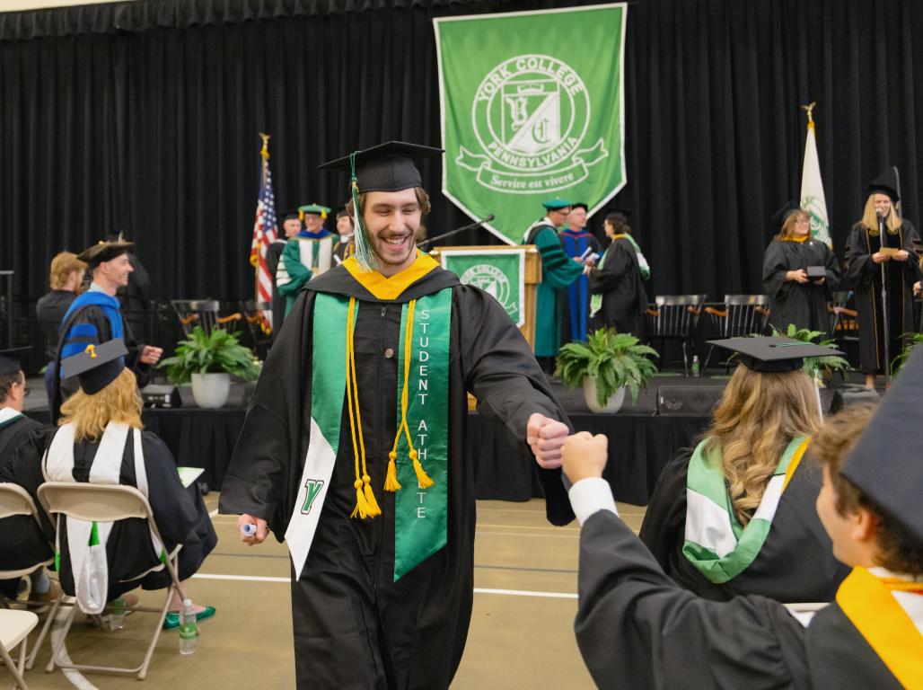 Students in cap and gowns for commencement. Student fist bumps another student with the stage behind him.
