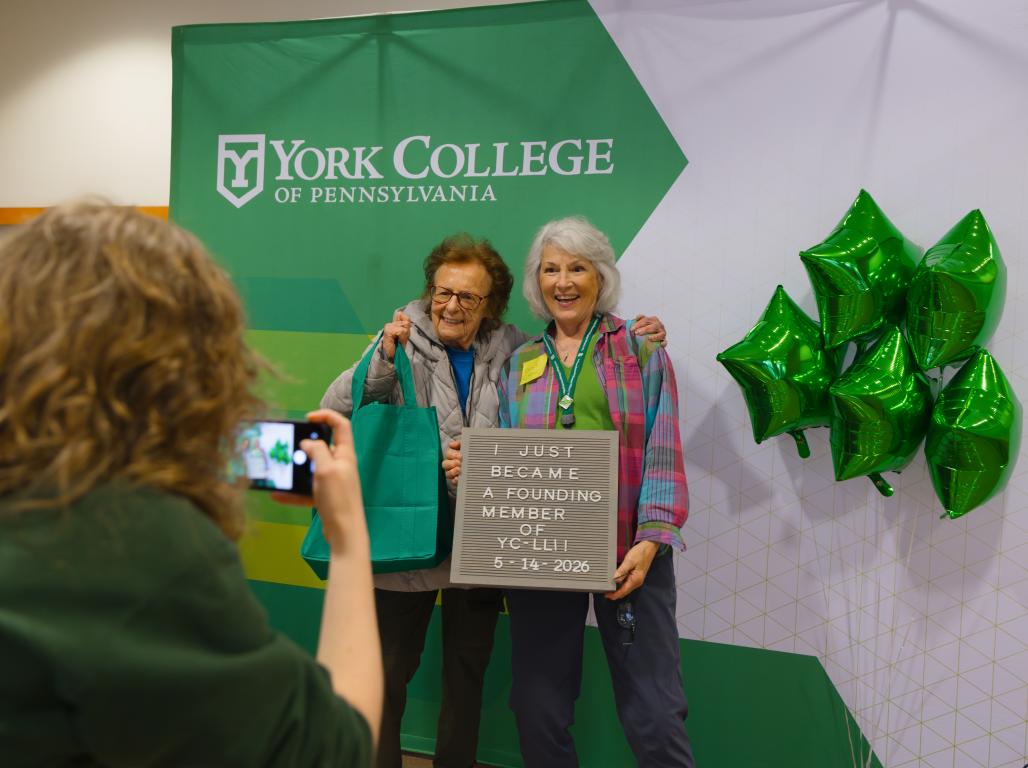Two women pose in front of a York College banner holding a sign that reads , "I just became a founding member of YC-LLI"