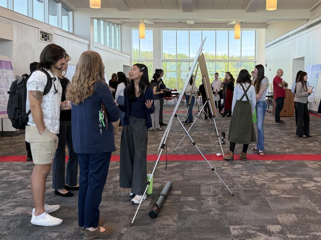 A group of students with poster presentations in a large room with tall windows.
