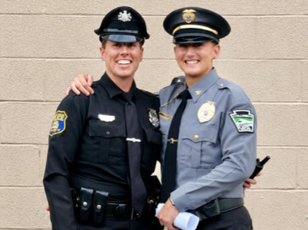 Two uniformed police officers smile with their arms around each other against a light-colored brick wall, while a police dog wearing a small officer’s cap sits in the foreground.