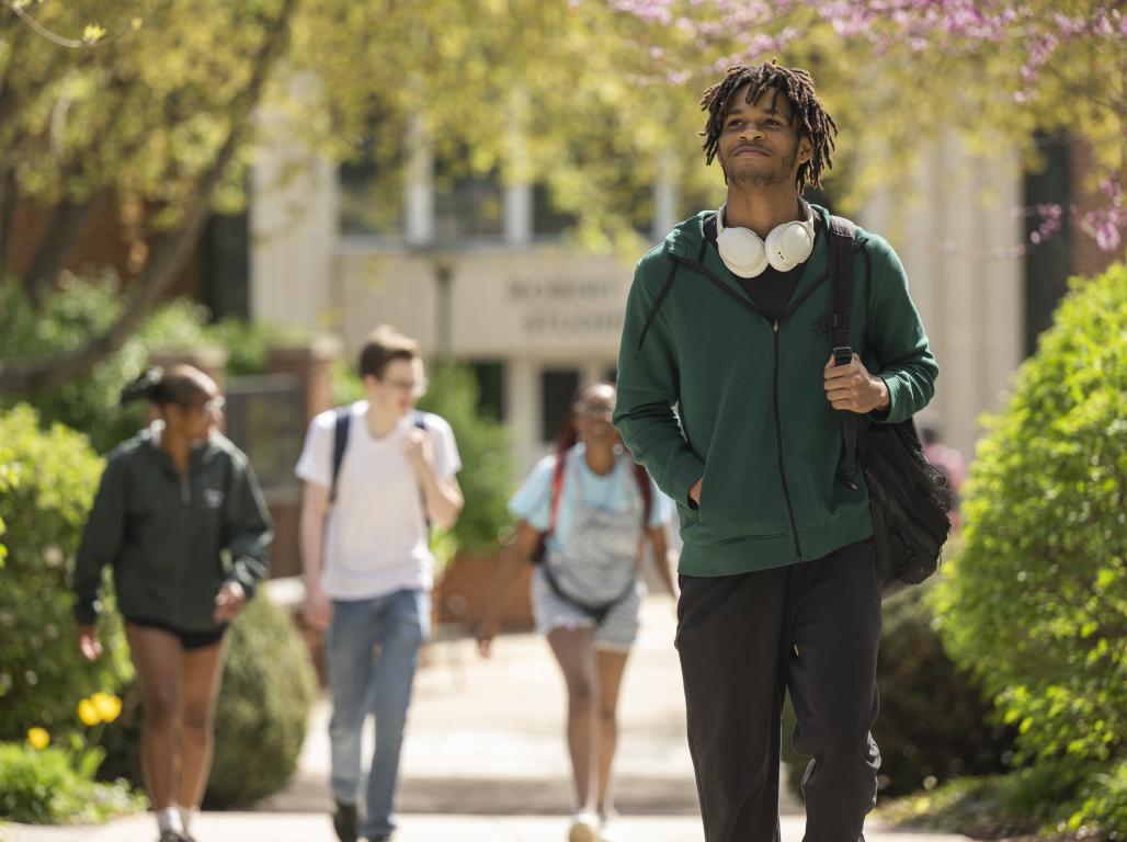 A college student with headphones and a backpack walks through a sunny campus courtyard.