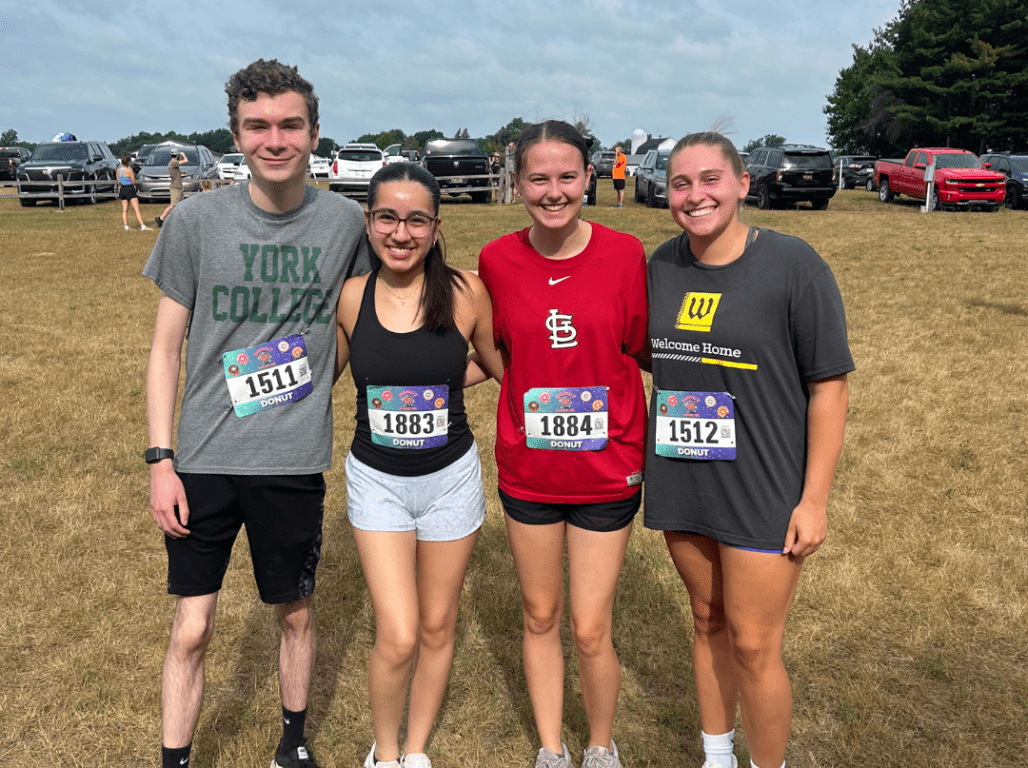 Four students smile together outdoors while wearing race bibs, with parked cars and trees in the background.