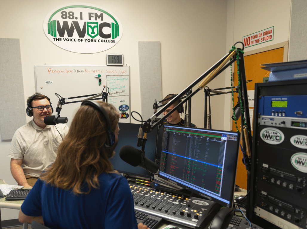 Three York College students wearing headphones broadcast from the WVYC radio studio beneath the 88.1 FM WVYC sign.
