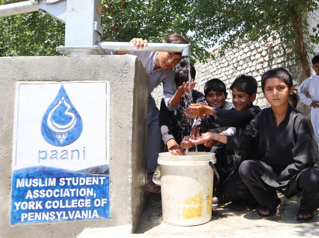 A group of children gather around a Paani Project well in Pakistan, using the hand pump and collecting water in a bucket.