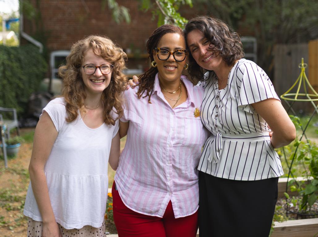 Three women stand together smiling in an outdoor community garden, with raised garden beds and greenery behind them.