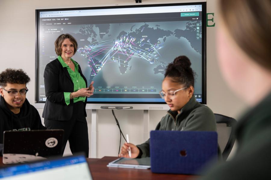 Dr. Tamara Schwartz instructing a cybersecurity class with a cyberthreat map while students take notes in the foreground.
