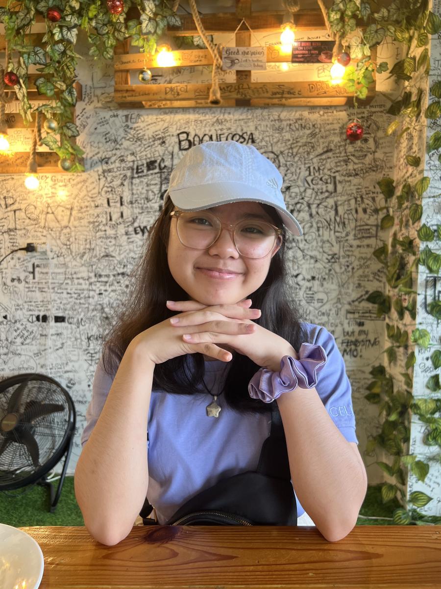 Person wearing a tan hat and purple shirt smiles at the camera in a cafe setting.