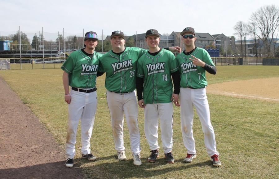 Four York College athletes in green and white uniforms smiling and posing on a baseball field.