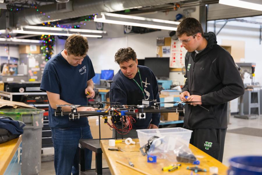 Three people looking intently at a drone on a workbench in an engineering lab setting.