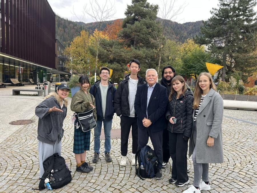 A group of people smile at the camera, they are outdoors on what appears to be a college campus.