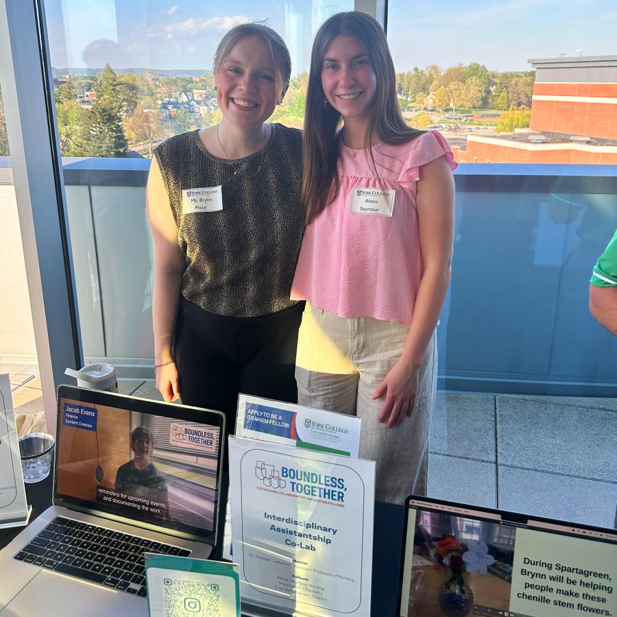 Two women stand together in front of a window with trees behind them. A laptop is open in front of them.