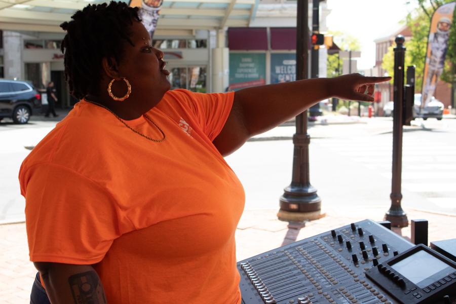 Person wearing an orange shirt standing at a sound board at an outdoor event. They are pointing to something out of frame.