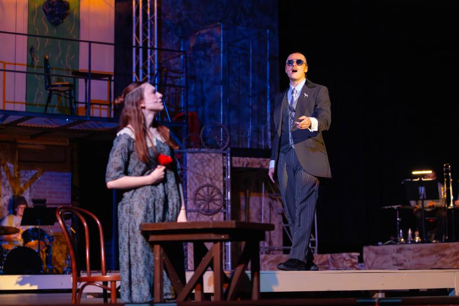 A person in a suit and sunglasses stands on a stage, gesturing while another person holds a red rose, facing them. The background includes stage props such as a table, chairs, and various set decorations.