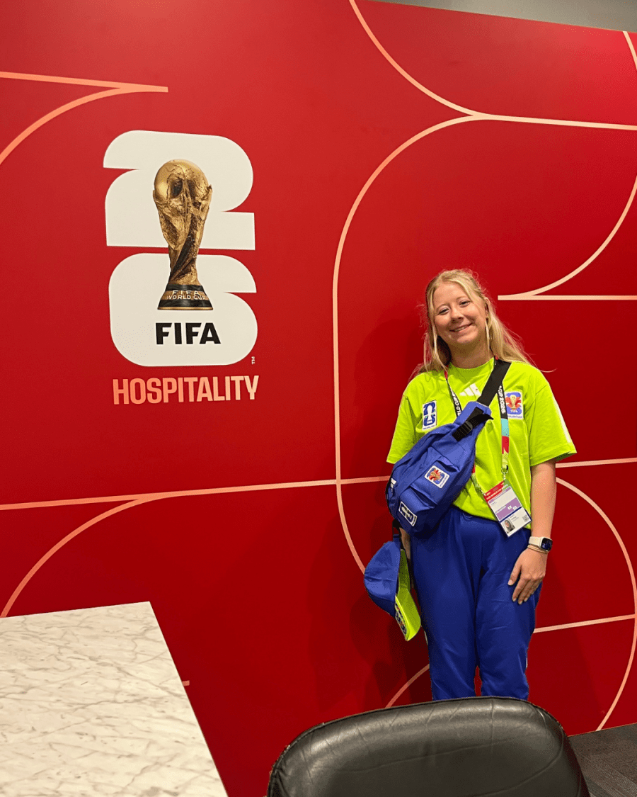Haley Feist ’26 stands in volunteer apparel beside a red FIFA Hospitality wall with a World Cup trophy graphic and event credential visible.