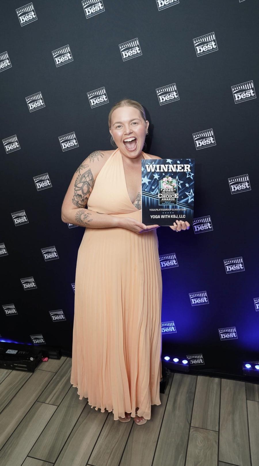 Kirsten Blair-Johnson ’18 smiles while holding a 2026 Community Choice Awards winner plaque for Yoga with KBJ, LLC in front of a Best of the Best backdrop.