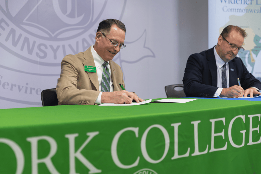 Two representatives sign documents at a York College partnership ceremony, seated behind a green York College tablecloth with Widener Law Commonwealth signage in the background.