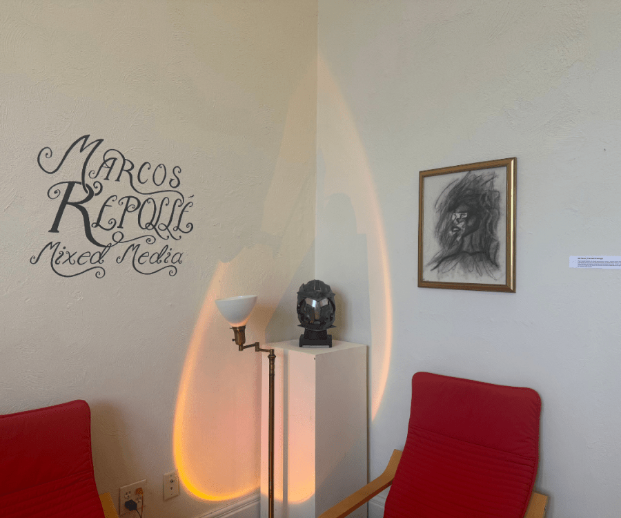 A Marketview Arts exhibit corner features wall text for a mixed media display, a framed charcoal-style portrait, a sculptural helmet on a pedestal, warm lighting, and red chairs.