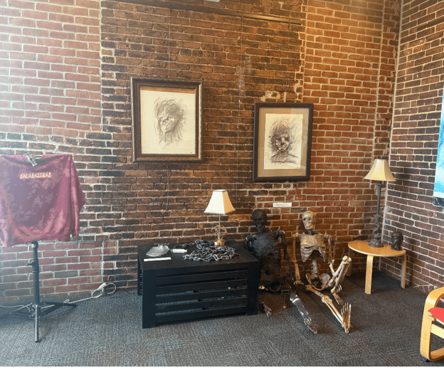 A Marketview Arts exhibit corner features wall text for a mixed media display, a framed charcoal-style portrait, a sculptural helmet on a pedestal, warm lighting, and red chairs.